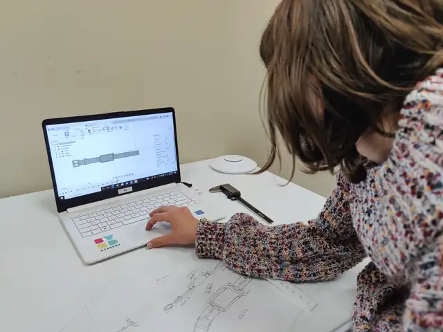 A student designs a part on a laptop beside her sketch